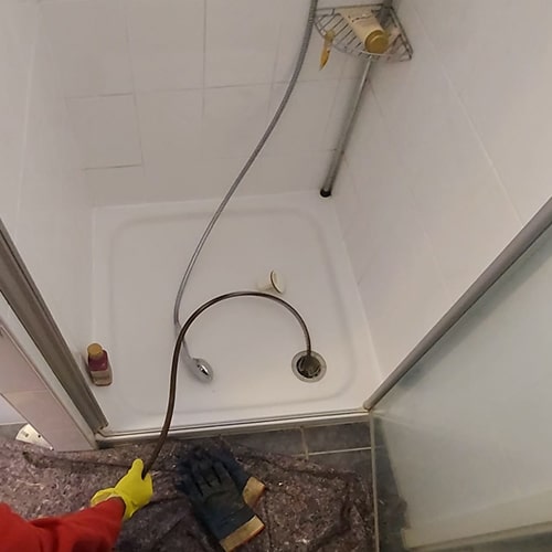 Rohrreinigung in einer Dusche: Person mit Handschuhen führt eine Rohrreinigungsspirale in den Abfluss ein.